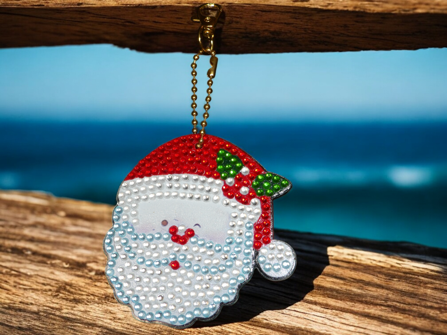Santa Clause Face Keychain with red hat and pearl-like beads hanging on a wooden surface against a blue background